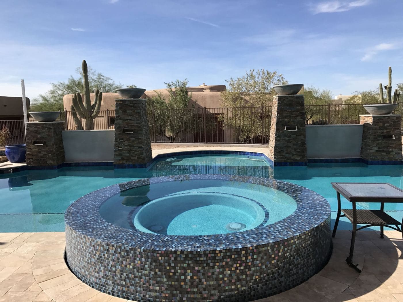 Raised round spa with mosaic tile beside a pool, stone pillars and saguaro cactus